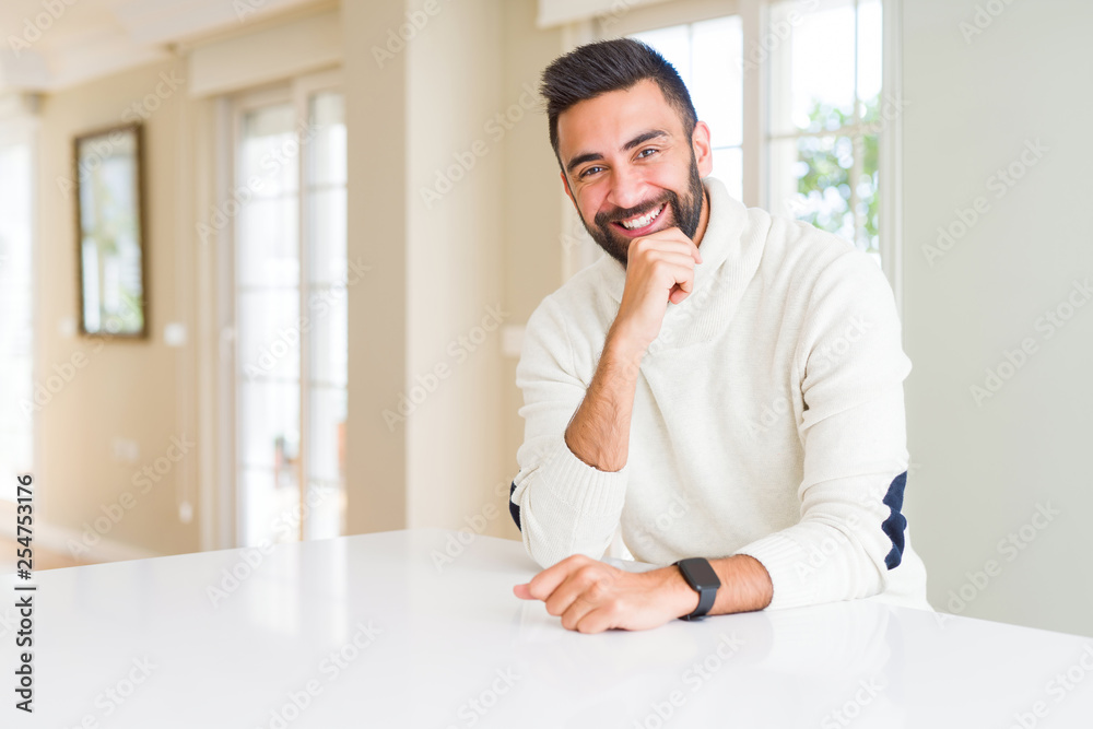 Handsome hispanic man wearing casual white sweater at home looking confident at the camera with smile with crossed arms and hand raised on chin. Thinking positive.