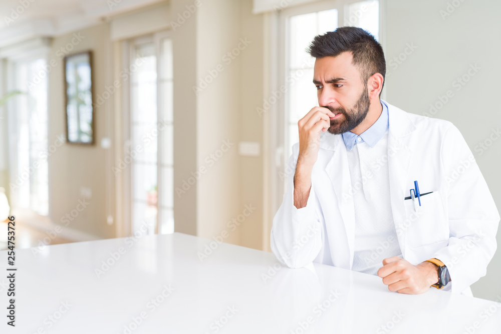Handsome hispanic doctor or therapist man wearing medical coat at the clinic looking stressed and nervous with hands on mouth biting nails. Anxiety problem. Handsome hispanic doctor or therapist man wearing medical coat at the clinic looking stressed and nervous with hands on mouth biting nails. Anxiety problem.