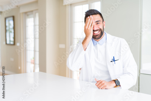 Handsome hispanic doctor or therapist man wearing medical coat at the clinic covering one eye with hand with confident smile on face and surprise emotion.