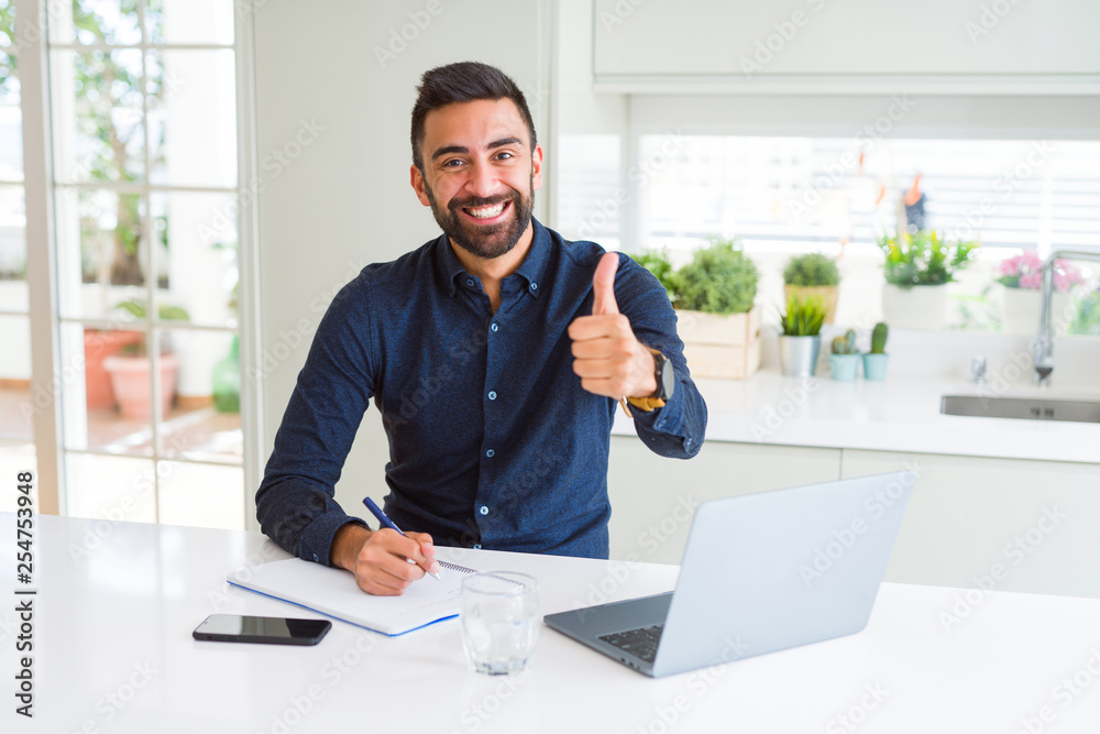 Handsome hispanic man working using computer and writing on a paper happy with big smile doing ok sign, thumb up with fingers, excellent sign