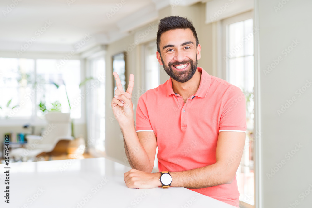 Handsome hispanic man wearing casual t-shirt at home smiling with happy face winking at the camera doing victory sign. Number two.