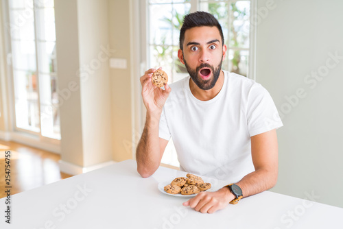 Handsome hispanic man eating chocolate chips cookies scared in shock with a surprise face  afraid and excited with fear expression