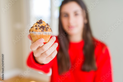 Beautiful young woman eating chocolate chips muffin with a confident expression on smart face thinking serious