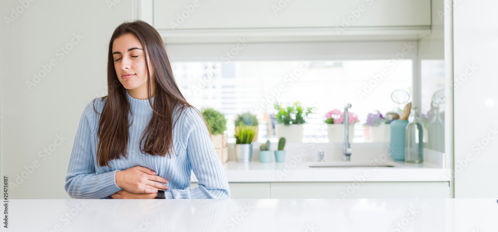 Wide angle picture of beautiful young woman sitting on white table at home with hand on stomach because nausea, painful disease feeling unwell. Ache concept. Wide angle picture of beautiful young woman sitting on white table at home with hand on stomach because nausea, painful disease feeling unwell. Ache concept.