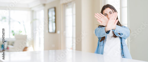 Wide angle picture of beautiful young woman sitting on white table at home Rejection expression crossing arms and palms doing negative sign, angry face