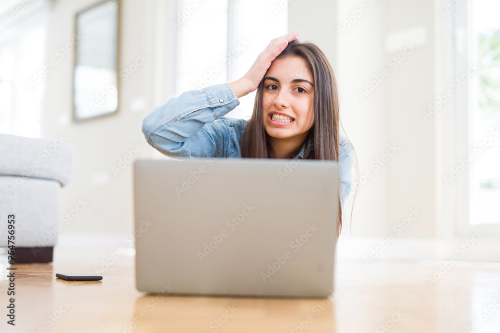Fototapeta premium Beautiful young woman laying on the floor using laptop stressed with hand on head, shocked with shame and surprise face, angry and frustrated. Fear and upset for mistake.