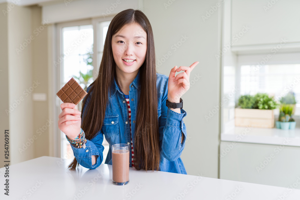 Beautiful Asian woman drinking chocolate milkshake and holding chocolate bar very happy pointing with hand and finger to the side Beautiful Asian woman drinking chocolate milkshake and holding chocolate bar very happy pointing with hand and finger to the side