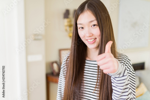 Beautiful Asian woman wearing stripes sweater over living room background doing happy thumbs up gesture with hand. Approving expression looking at the camera showing success.