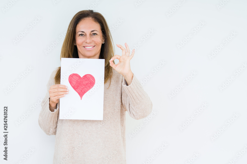 Middle age woman holding card gift with red heart over isolated background doing ok sign with fingers, excellent symbol Middle age woman holding card gift with red heart over isolated background doing ok sign with fingers, excellent symbol