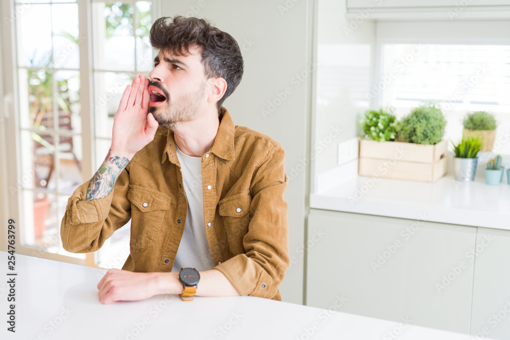 Young man wearing casual jacket sitting on white table shouting and screaming loud to side with hand on mouth. Communication concept. Young man wearing casual jacket sitting on white table shouting and screaming loud to side with hand on mouth. Communication concept.