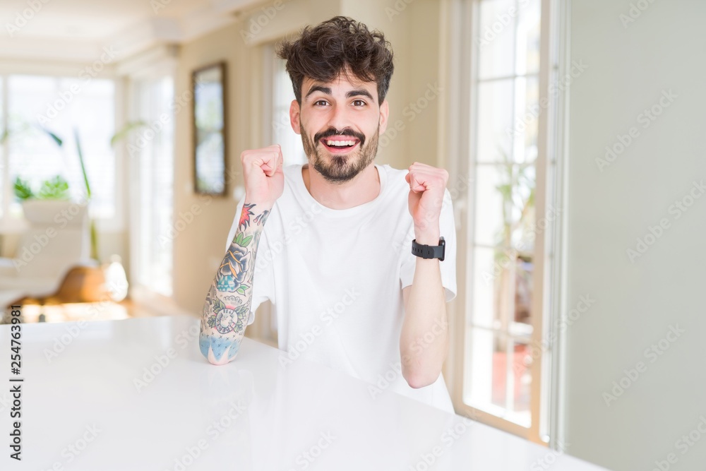Young man wearing casual t-shirt sitting on white table celebrating surprised and amazed for success with arms raised and open eyes. Winner concept. Young man wearing casual t-shirt sitting on white table celebrating surprised and amazed for success with arms raised and open eyes. Winner concept.