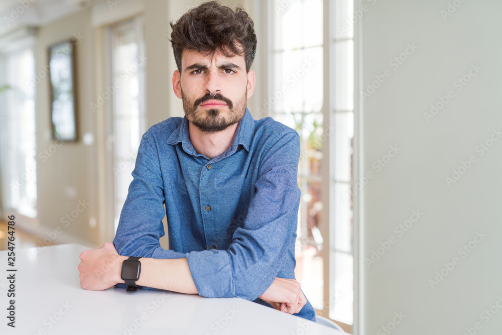 Young man wearing casual shirt sitting on white table skeptic and nervous, disapproving expression on face with crossed arms. Negative person.
