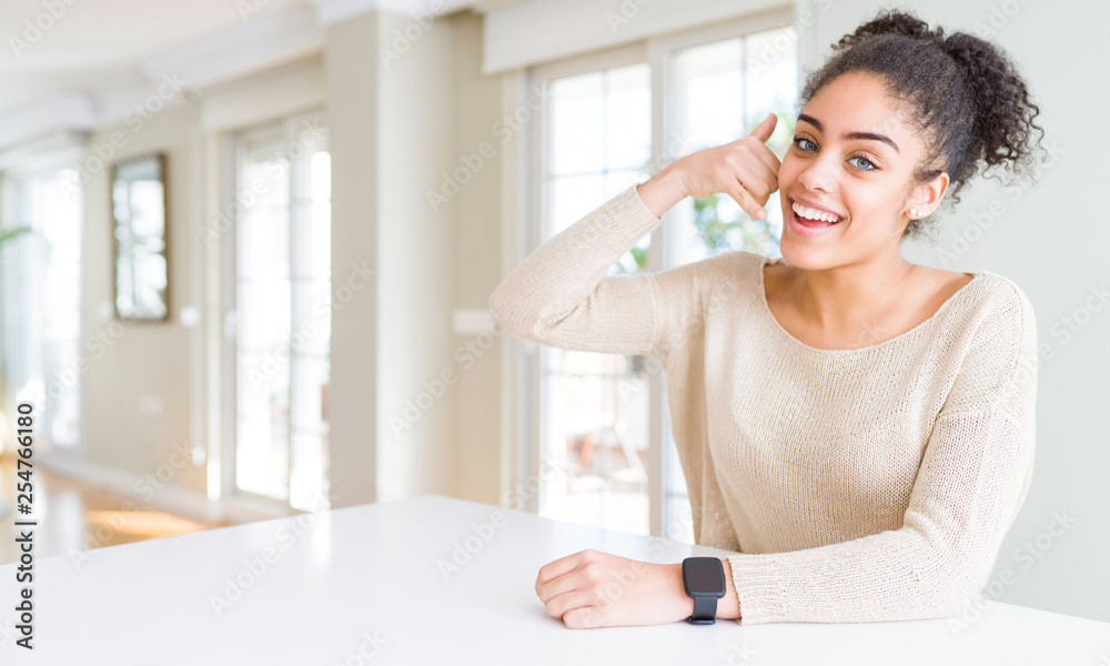 Beautiful young african american woman with afro hair sitting on table at home smiling doing phone gesture with hand and fingers like talking on the telephone. Communicating concepts. Beautiful young african american woman with afro hair sitting on table at home smiling doing phone gesture with hand and fingers like talking on the telephone. Communicating concepts.