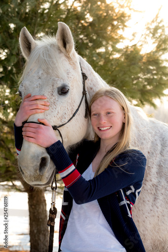 happy blonde teenager girl holding white/grey handsome horse. Sun is back lighting the scene and both are looking toward camera. 