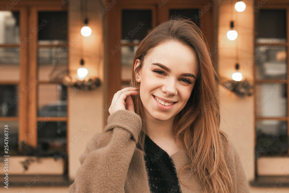Close up portrait of a happy girl in warm spring clothes is standing on the street in the background of a brown wall with windows, looking at the camera and smiling.Cute girl in a coat on the street Close up portrait of a happy girl in warm spring clothes is standing on the street in the background of a brown wall with windows, looking at the camera and smiling.Cute girl in a coat on the street