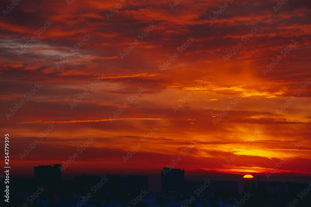 Cityscape with vivid fiery dawn. Amazing warm dramatic cloudy sky above dark silhouettes of city buildings. Orange sunlight. Atmospheric background of sunrise in overcast weather. Copy space.