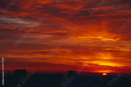 Cityscape with vivid fiery dawn. Amazing warm dramatic cloudy sky above dark silhouettes of city buildings. Orange sunlight. Atmospheric background of sunrise in overcast weather. Copy space.