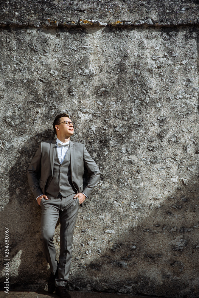 Portrait of a modern handsome guy, in formal stylish clothes, who is posing against the gray wall outdoors. Attractive groom
