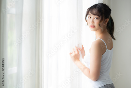 Portrait of beautiful young asian woman standing at the window and smile while wake up with sunlight at morning, girl happy with fresh and cheerful, lifestyle and relax for wellness concept.