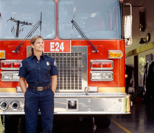 Smiling female woman firefighter in uniform with fire engine truck at station. Happy, confident, successful public service career professional.
