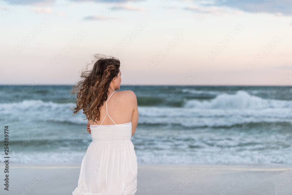 Young woman back standing in white dress on beach evening in Florida panhandle shivering arms crossed in wind by ocean waves