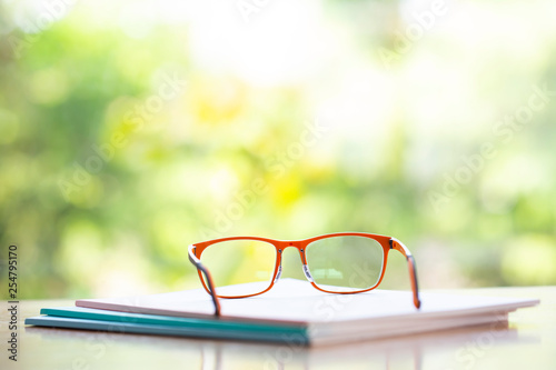 Orange eyeglasses with three white notebook on wooden table, Bokeh garden background, Close up & Macro shot, Selective focus, Stationery concept