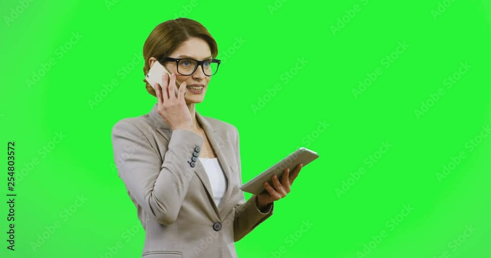 Caucasian young good looking woman in glasses and business style talking on the mobile phone and holding a tablet device on the green background. Alpha channel. Caucasian young good looking woman in glasses and business style talking on the mobile phone and holding a tablet device on the green background. Alpha channel.