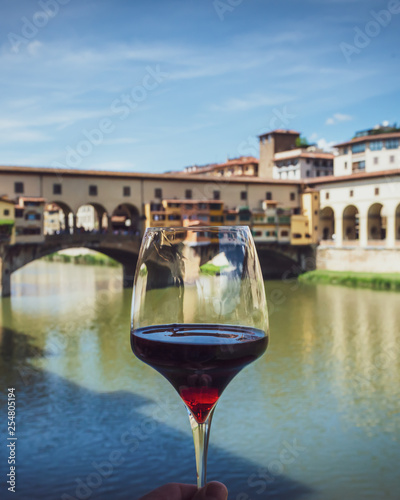 Glass of red wine on background of city and famous bridge in Florence, Italy. View from restaurant terrace on antique buildings in warm summer day.