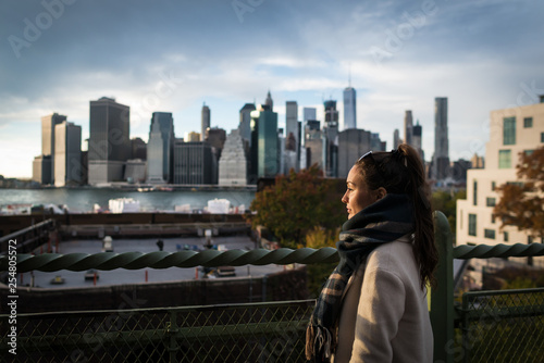 Young woman standing on riverside in Brooklyn and looking on Manhattan skyscrapers. Elegant girl on background of city downtown in New York, USA.