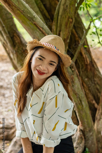 Women white skin lovely brown hair wearing a basketry hat brown red lip wear white shirt wearing black pants women sit poses photography portrait under the tree In the garden.