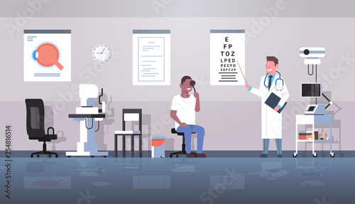 male ophthalmologist checking african american man patient eyesight doctor in uniform pointing letters at eye chart medicine and healthcare concept oculists office interior horizontal