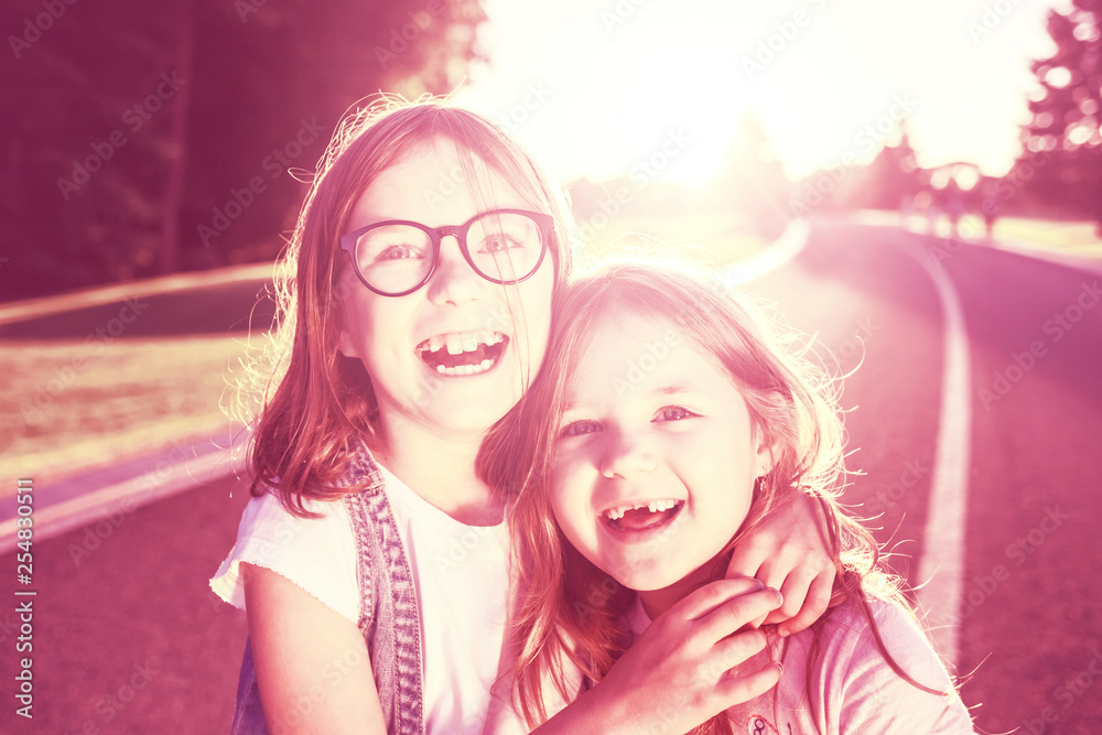 Two joyful girls standing on the road, hugging and smiling against the sunset. Girls without front teeth. Toned in retro styled colors. Two joyful girls standing on the road, hugging and smiling against the sunset. Girls without front teeth. Toned in retro styled colors.