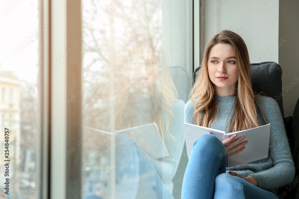Beautiful young woman reading book while sitting on window sill Beautiful young woman reading book while sitting on window sill