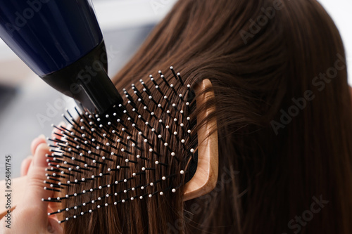 Hairdresser with blow drier brushing long hair of young woman in salon, closeup