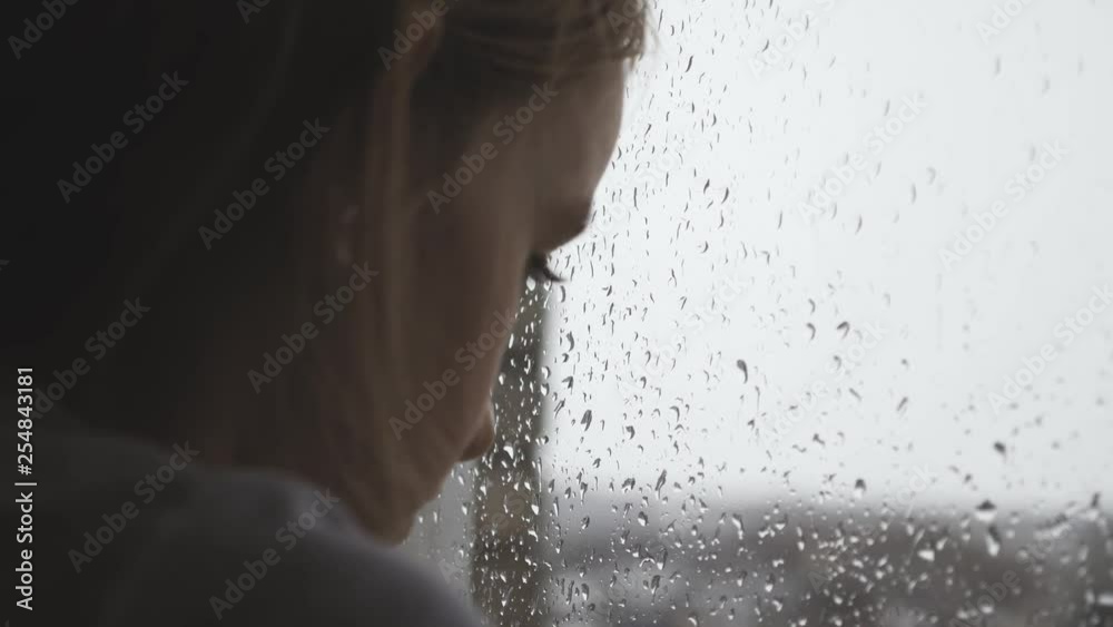 Sad teenager girl looking outside through the window in the rainy day.