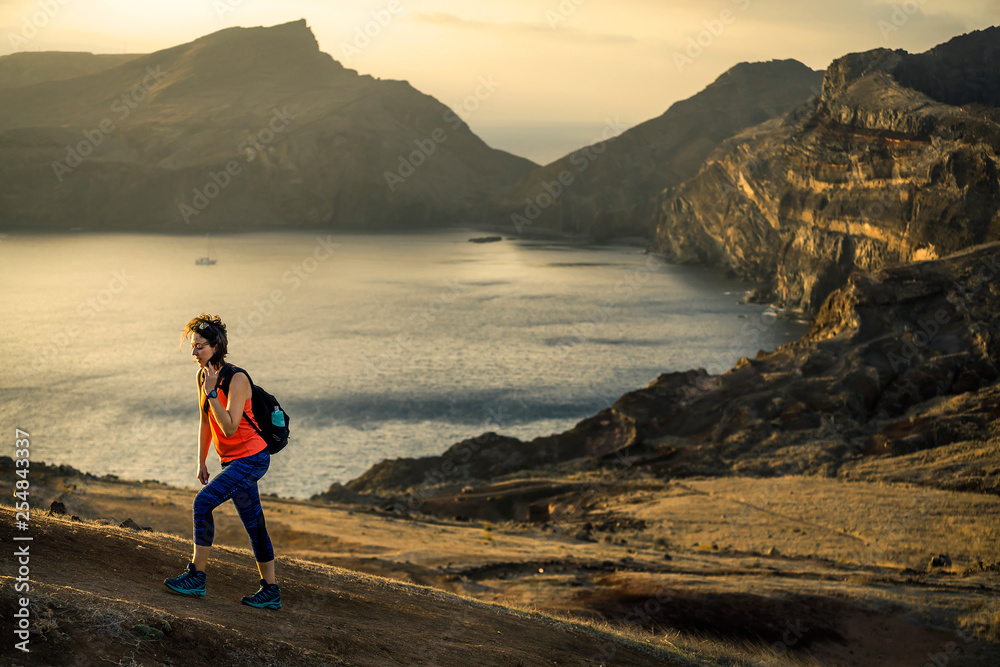 Young and attractive girl in orange shirt and blue leggings is hiking by the tourist's trail on Madeira island, Portugal. Sunny day. The end of the trail, she is climbing up on sunset.