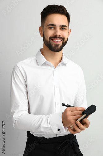 Handsome waiter with notebook on light background