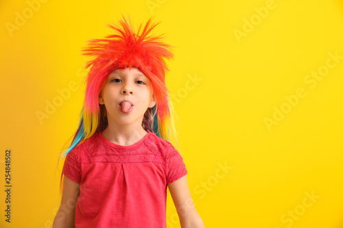 Funny little girl in wig showing tongue on color background. April fools' day celebration