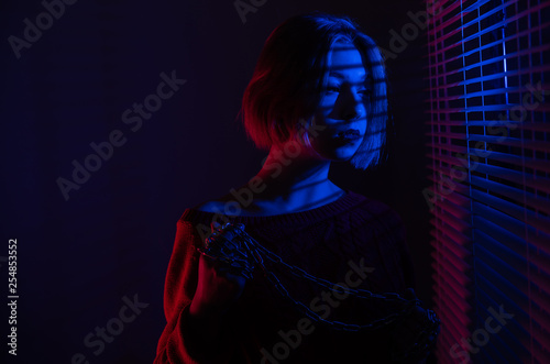 A beautiful girl with a chain in her hands stands near the window with blinds in the light of neon lights of blue and red light