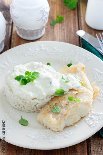 Fried cod, cooked with rice and cream sauce on a white plate, selective focus