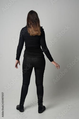 Fotografía full length portrait of a brunette girl wearing  modern black jacket and pants, standing pose with back to the camera on grey studio background