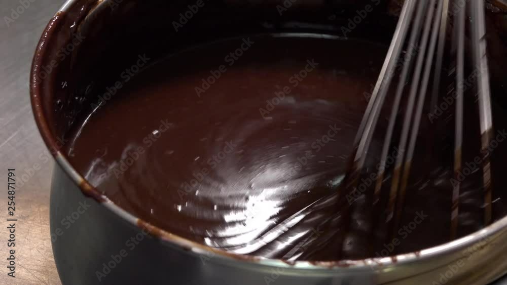 A baker stirs liquid chocolate frosting in a metallic bowl with a whisk - closeup A baker stirs liquid chocolate frosting in a metallic bowl with a whisk - closeup