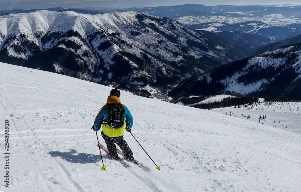 Male skier skiing freeride downhill with view to valley of Low Tatras, Slovakia. Colorful jacket with black backpack