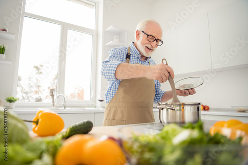 Close up photo grey haired he his him grandpa appetite waiting guests cooking favorite family dish mix all in pan ready wear specs casual checkered plaid shirt jeans denim outfit kitchen indoors