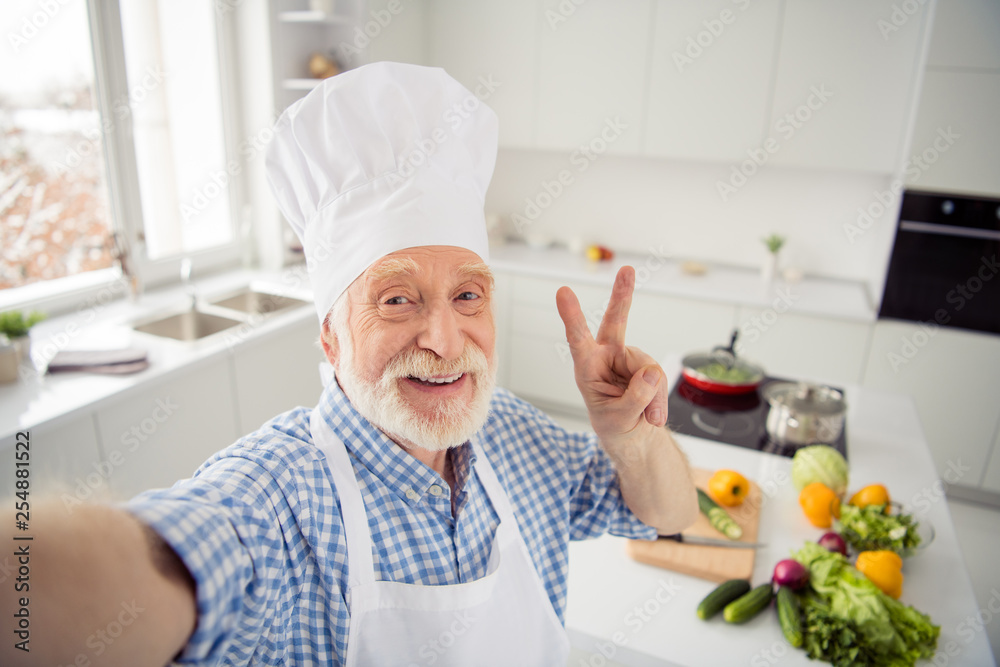 Close up photo cheer grey haired he his him grandpa telephone make take selfies video followers show v-sign say hi wear baker chefs costume casual checkered plaid shirt outfit house kitchen Close up photo cheer grey haired he his him grandpa telephone make take selfies video followers show v-sign say hi wear baker chefs costume casual checkered plaid shirt outfit house kitchen
