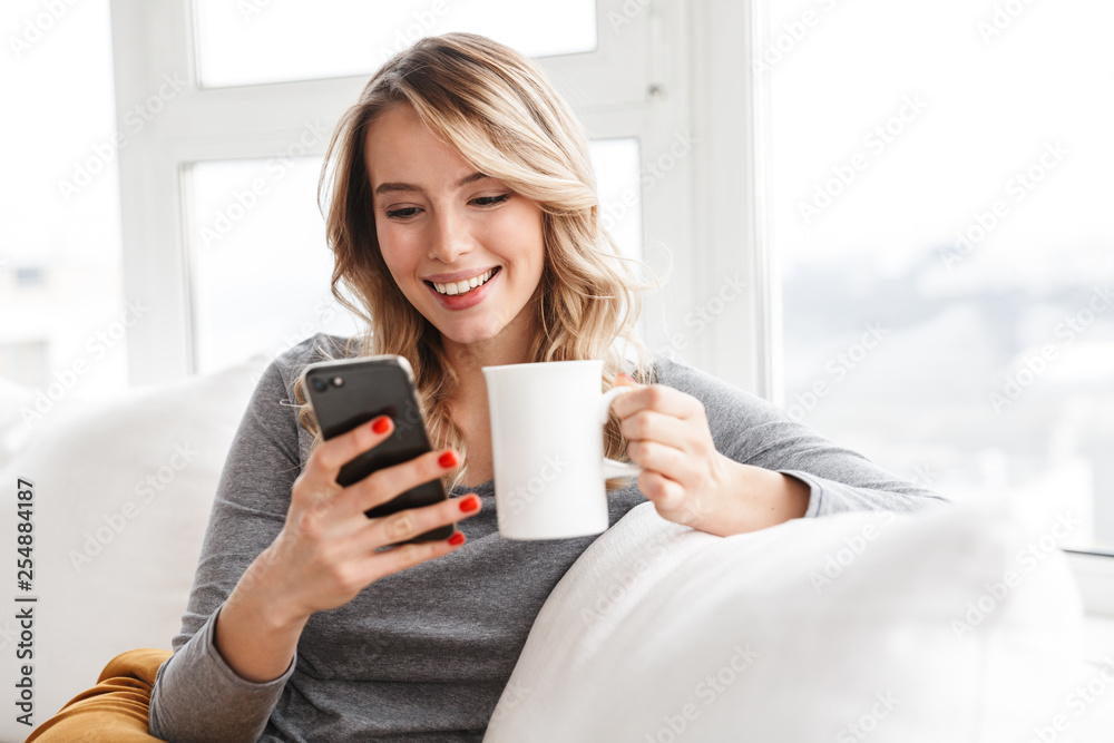 Pretty woman sitting indoors at home holding cup drinking tea using mobile phone. Pretty woman sitting indoors at home holding cup drinking tea using mobile phone.