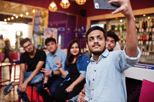 Group of stylish asian friends wear on jeans sitting at chairs against bar in club and makes selfie together.