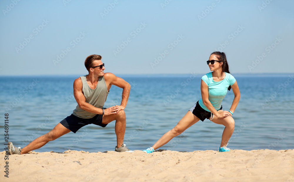fitness, sport and lifestyle concept - smiling couple stretching legs on beach before training fitness, sport and lifestyle concept - smiling couple stretching legs on beach before training