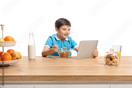 Little boy eating cereals for brekfast at a wooden counter and watching in a tablet