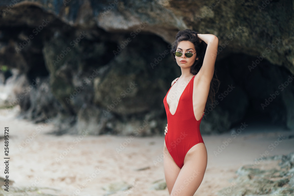 Slender tanned girl in swimwear and sunglasses posing near stone at rocky beach by ocean.